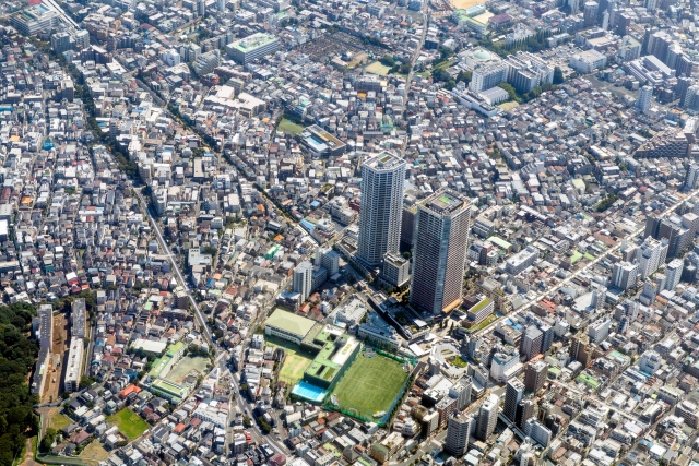 Aerial view of a dense cityscape with two tall office towers and a green sports field nearby.
